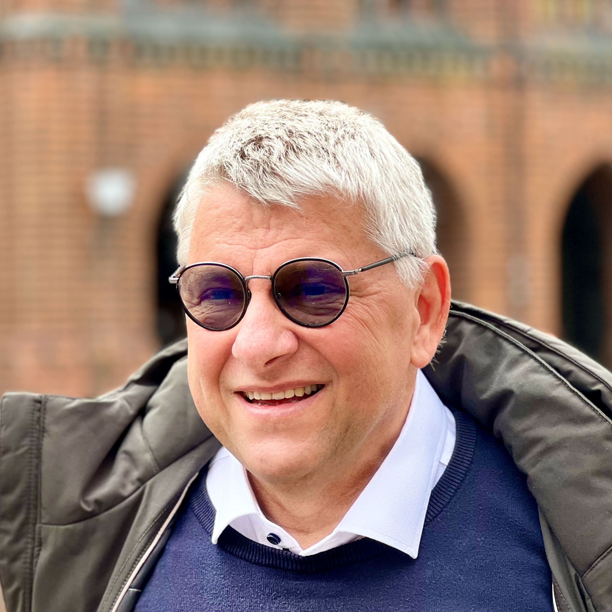 Lächelnder Mann mit grauen Haaren und Sonnenbrille vor Backsteingebäude.