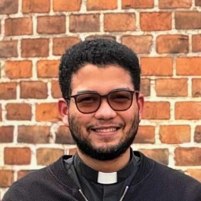 Dennis Porträt eines lächelnden Priesters mit Brille und Priesterkragen.