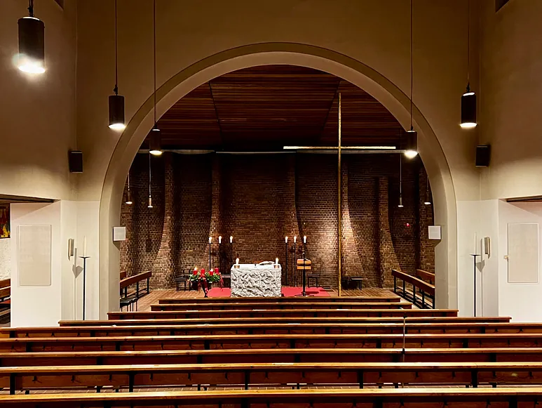 Kircheninnenraum mit Bänken, Steinaltar und großem Kreuz.