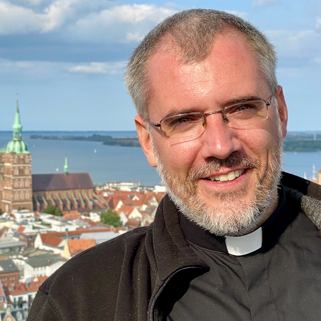 Lächelnder Priester mit der Stralsunder Altstadt im Hintergrund.