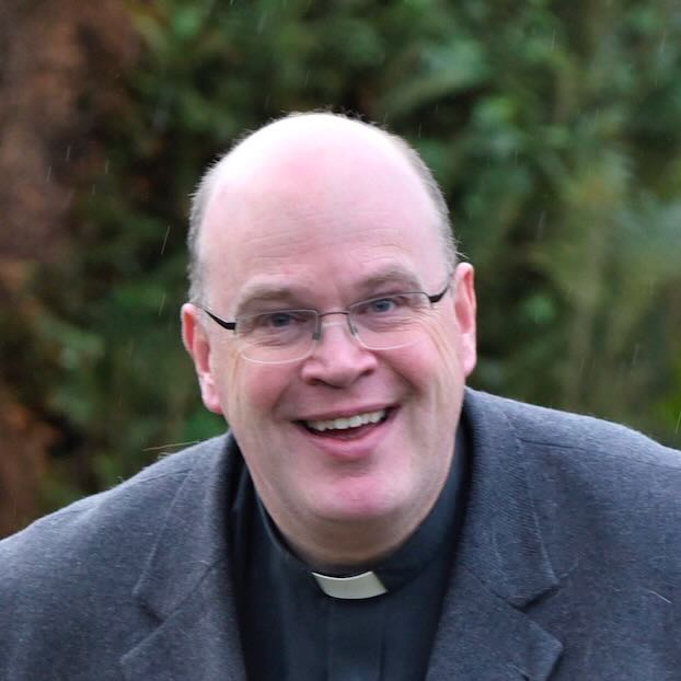 Porträt eines lächelnden Priesters mit Brille und Priesterkragen.